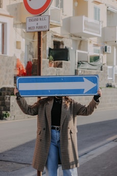 A person stands on a street holding up a large blue right arrow sign, with the sign covering their face. Behind the person, there is a traffic post with Italian text and a no-entry symbol. The background includes a rough stone wall and residential buildings with balconies.
