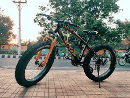 A black and orange mountain bike with thick tires is parked on a tiled sidewalk. It stands in front of a street with some trees visible in the background. The bike has the word 'VOLTEBIKE' written on its frame and appears to be brand new. There is a motorbike passing by in the background.