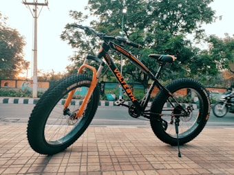 A black and orange mountain bike with thick tires is parked on a tiled sidewalk. It stands in front of a street with some trees visible in the background. The bike has the word 'VOLTEBIKE' written on its frame and appears to be brand new. There is a motorbike passing by in the background.