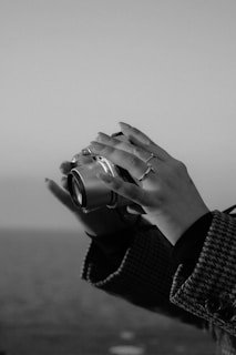 A monochrome image featuring a close-up of hands holding a camera. The hands are adorned with rings, and the person is wearing a houndstooth-patterned coat. The background appears blurred, hinting at an outdoor setting, possibly near water.