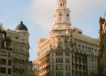 A cityscape featuring elegant and historic European architecture with multiple stories, large windows, and ornate details. Dominant is a tall, elegant building with a dome on the left side, set against a clear sky with soft clouds.
