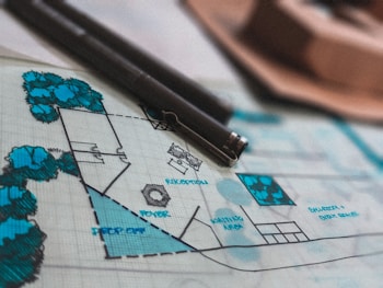 A detailed architectural blueprint lies on a table, adorned with various labels such as 'RECEPTION,' 'FOYER,' and 'IDENTIFICA AREA.' Several blue trees and shrubbery areas are illustrated on the plan. Two black pens rest on the blueprint, indicating ongoing work or recent annotations.