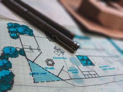 A detailed architectural blueprint lies on a table, adorned with various labels such as 'RECEPTION,' 'FOYER,' and 'IDENTIFICA AREA.' Several blue trees and shrubbery areas are illustrated on the plan. Two black pens rest on the blueprint, indicating ongoing work or recent annotations.