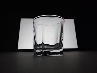 A clear, empty glass with a slightly reflective surface is positioned against a contrasting background of white and black. The lighting creates soft shadows and highlights on the glass surface, adding to its transparent and minimalistic look.