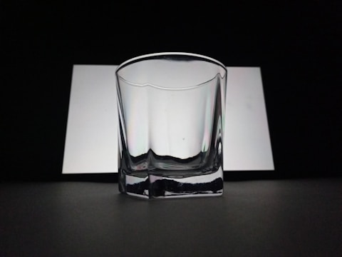 A clear, empty glass with a slightly reflective surface is positioned against a contrasting background of white and black. The lighting creates soft shadows and highlights on the glass surface, adding to its transparent and minimalistic look.