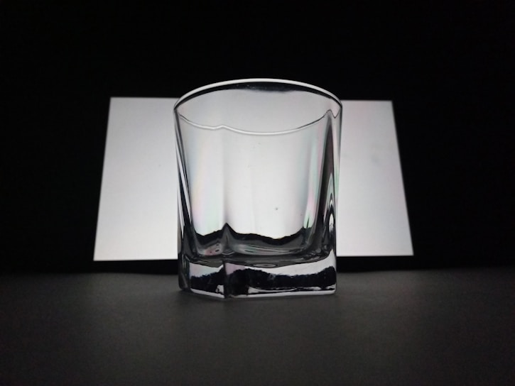 A clear, empty glass with a slightly reflective surface is positioned against a contrasting background of white and black. The lighting creates soft shadows and highlights on the glass surface, adding to its transparent and minimalistic look.