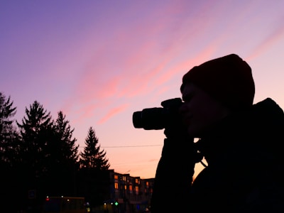 A silhouette of a person using a camera against a backdrop of a vibrant sunset sky with shades of pink and purple. In the background, tall pine trees and buildings with illuminated windows are visible, adding to the serene and picturesque scene.