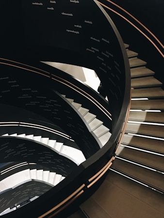 A spiral staircase with multiple levels and contrasting black and white colors. The bannisters are sleek and smooth, creating a modern and elegant appearance. Words in a foreign language are visible on the walls of the staircase, adding an intriguing element.