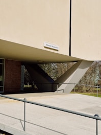 A modern architectural structure featuring large concrete supports and an elevated platform. A sign with the address 'Altonaer Strasse 4-14' is mounted on the building. The surrounding area includes a pathway guarded by railings and greenery with trees.