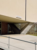 A modern architectural structure featuring large concrete supports and an elevated platform. A sign with the address 'Altonaer Strasse 4-14' is mounted on the building. The surrounding area includes a pathway guarded by railings and greenery with trees.