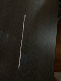 A delicate, sparkling bracelet made of small, evenly spaced stones is laid out on a dark wooden surface. The bracelet appears to be finely crafted and catches light subtly across its length.