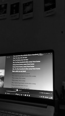 A monochrome photo displaying a laptop in a dimly lit room. The screen shows a music player with song lyrics of 'Five Hundred Miles' prominently displayed. Above the laptop, there are four polaroid-style pictures hanging on the wall.