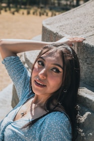 A woman with long dark hair leans against a stone wall with one arm raised. She is wearing a light blue patterned dress and smiling. The background is an outdoor setting with blurry green foliage.