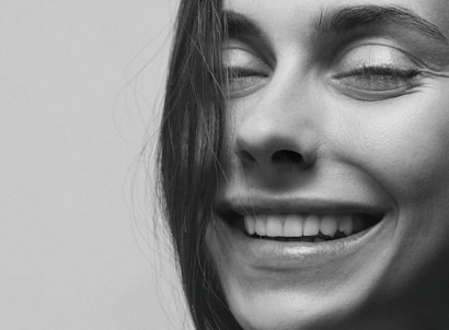 A close-up black and white photograph of a woman's face with a joyful expression, featuring a broad smile and eyes gently closed. A few strands of hair softly fall across the face.