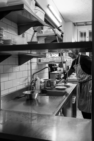 A black and white image depicting a person working in a kitchen setting. The individual is wearing a striped apron and is positioned near a stainless steel counter with a variety of cooking utensils. A faucet with a flexible hose hangs above the sink area, and there are shelves with containers and dishes above the counter. The setting appears clean and well-organized, suggesting a professional kitchen environment.