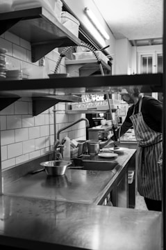 A black and white image depicting a person working in a kitchen setting. The individual is wearing a striped apron and is positioned near a stainless steel counter with a variety of cooking utensils. A faucet with a flexible hose hangs above the sink area, and there are shelves with containers and dishes above the counter. The setting appears clean and well-organized, suggesting a professional kitchen environment.