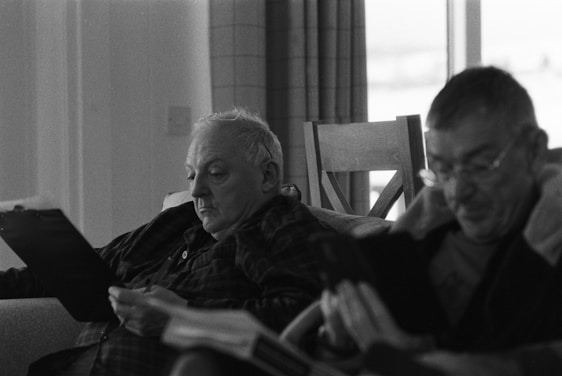 Two people are seated indoors, each focused on reading material, possibly a book or document. The setting has a relaxed atmosphere, with soft lighting and a visible chair in the background.
