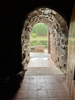 A stone archway leading to an outdoor setting with a view of a forest and a small body of water. The passageway has a textured stone interior and opens onto a tiled floor outside. The scene beyond the archway features green foliage and a tranquil pond.