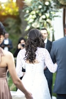 A bride with long, dark curly hair is seen from behind, wearing a lace-up white wedding dress with intricate lace details. She is accompanied by a man in a grey suit and another woman in a bronze dress. The background includes a floral arrangement with white and green flowers, and blurred figures suggesting a wedding ceremony setting.