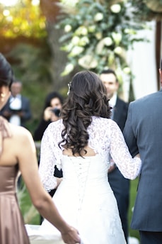 A bride with long, dark curly hair is seen from behind, wearing a lace-up white wedding dress with intricate lace details. She is accompanied by a man in a grey suit and another woman in a bronze dress. The background includes a floral arrangement with white and green flowers, and blurred figures suggesting a wedding ceremony setting.