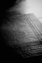 A black-and-white image featuring the textured surface of a book cover with an embossed design. The design includes geometric patterns and symbolic imagery, and the light highlights the fine details of the cover material.