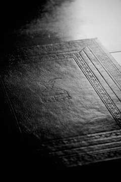 A black-and-white image featuring the textured surface of a book cover with an embossed design. The design includes geometric patterns and symbolic imagery, and the light highlights the fine details of the cover material.
