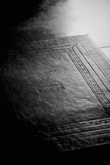 A black-and-white image featuring the textured surface of a book cover with an embossed design. The design includes geometric patterns and symbolic imagery, and the light highlights the fine details of the cover material.