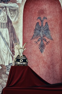 A mural or fresco depicting a stylized double-headed eagle is set against a red background, with artistic detailing on its feathers and feet. To the left, there is a partial view of a historical or mythical figure in detailed attire. In the foreground, a sculptural piece resembling a goat's head, adorned with decorative elements resembling a crown or armor, is placed on a table draped with deep red fabric.