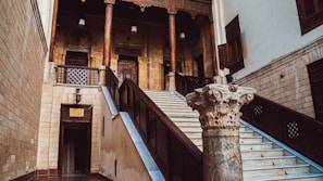 An ornate staircase leads up to a set of wooden doors framed by intricate architectural details. A single stone column with elaborate carvings stands prominently in the foreground. The walls are constructed from light-colored stone, and the structure features wooden elements and traditional design, lending an air of historical significance.