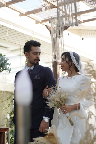 A man and woman in wedding attire stand together in a bright, airy environment with natural elements. The woman is holding a bouquet of dried flowers and wears a veil. The man is dressed in a dark suit and they appear to be exchanging a loving look.