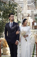 A couple stands under a decorated pergola adorned with greenery and cascading macrame chandeliers. The man is wearing a navy blue suit with a patterned tie, while the woman dons a flowing white dress and holds a dried flower bouquet. Elegant lighting fixtures hang above them, casting a warm atmosphere. The setting appears to be an outdoor celebration with a rustic and romantic theme.