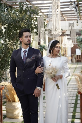 A couple stands under a decorated pergola adorned with greenery and cascading macrame chandeliers. The man is wearing a navy blue suit with a patterned tie, while the woman dons a flowing white dress and holds a dried flower bouquet. Elegant lighting fixtures hang above them, casting a warm atmosphere. The setting appears to be an outdoor celebration with a rustic and romantic theme.