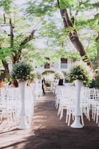 An outdoor wedding ceremony setup with white chairs arranged in rows facing a floral altar. Tall arrangements of greenery and white flowers are placed along the aisle. The setting is beneath lush green trees and appears to be part of a garden or courtyard.