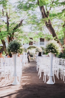 An outdoor wedding ceremony setup with white chairs arranged in rows facing a floral altar. Tall arrangements of greenery and white flowers are placed along the aisle. The setting is beneath lush green trees and appears to be part of a garden or courtyard.