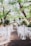An outdoor wedding ceremony setup with white chairs arranged in rows facing a floral altar. Tall arrangements of greenery and white flowers are placed along the aisle. The setting is beneath lush green trees and appears to be part of a garden or courtyard.