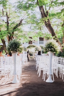 An outdoor wedding ceremony setup with white chairs arranged in rows facing a floral altar. Tall arrangements of greenery and white flowers are placed along the aisle. The setting is beneath lush green trees and appears to be part of a garden or courtyard.