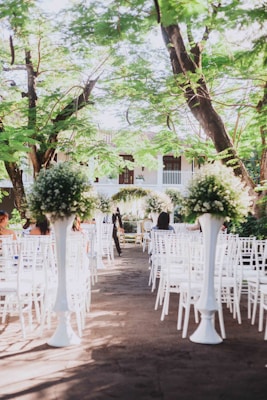 An outdoor wedding ceremony setup with white chairs arranged in rows facing a floral altar. Tall arrangements of greenery and white flowers are placed along the aisle. The setting is beneath lush green trees and appears to be part of a garden or courtyard.