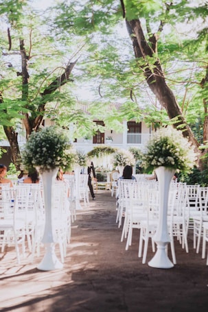 An outdoor wedding ceremony setup with white chairs arranged in rows facing a floral altar. Tall arrangements of greenery and white flowers are placed along the aisle. The setting is beneath lush green trees and appears to be part of a garden or courtyard.