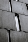 Several large, rectangular concrete panels are arranged in a grid pattern on the facade of a modern building. The panels have a textured surface and are separated by narrow, vertical window slits.