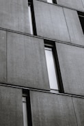 Several large, rectangular concrete panels are arranged in a grid pattern on the facade of a modern building. The panels have a textured surface and are separated by narrow, vertical window slits.