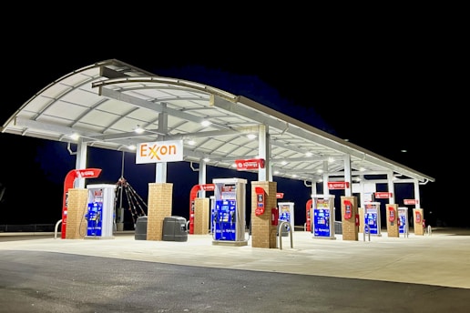 A well-lit gas station at night with multiple fuel pumps under a large canopy. The overhead structure is modern, featuring a slightly curved roof. The bright lighting highlights the cleanliness and newness of the station, with a prominent Exxon sign displayed above.