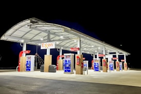 A well-lit gas station at night with multiple fuel pumps under a large canopy. The overhead structure is modern, featuring a slightly curved roof. The bright lighting highlights the cleanliness and newness of the station, with a prominent Exxon sign displayed above.