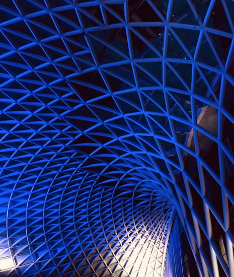 An intricate geometric structure made up of interlocking triangular shapes, with a gradient of blue lighting creating a dramatic effect. This architectural feature is reminiscent of a modern atrium or roof design, emphasizing depth and perspective.