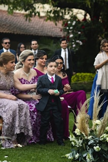 A group of people dressed in formal attire is gathered outdoors, likely at a wedding or formal event. A young boy in a suit stands in the foreground, surrounded by women in elegant dresses. The background features lush greenery and floral arrangements, creating a festive and celebratory atmosphere.