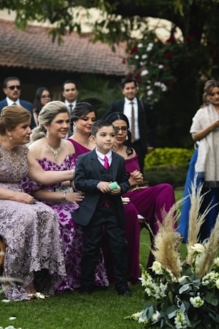 A group of people dressed in formal attire is gathered outdoors, likely at a wedding or formal event. A young boy in a suit stands in the foreground, surrounded by women in elegant dresses. The background features lush greenery and floral arrangements, creating a festive and celebratory atmosphere.