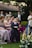 A group of people dressed in formal attire is gathered outdoors, likely at a wedding or formal event. A young boy in a suit stands in the foreground, surrounded by women in elegant dresses. The background features lush greenery and floral arrangements, creating a festive and celebratory atmosphere.
