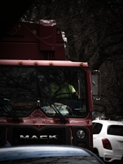 A red Mack truck is positioned on a street with bare tree branches in the background. The driver, wearing a yellow vest, is visible through the windshield. Nearby, a white and a blue car can be seen in close proximity.
