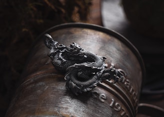 A detailed metal dragon sculpture is placed on an old, weathered barrel or container. The dragon is intricately designed with visible scales and flowing elements, lying atop the curved surface, which has faintly visible text inscribed on it. The background is dark and out of focus, emphasizing the dragon's craftsmanship.
