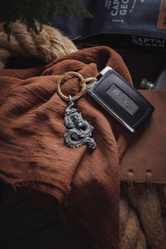 A car key with a metallic logo and a detailed dragon keychain lie on a textured brown fabric. In the background, a large rope and a leather-bound notebook are partially visible, with a package labeled 'Captain George' in the top right corner.