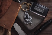 A collection of personal items arranged on a wooden surface, including a leather-bound notebook, a metal keychain with a dragon design, a car key for a Jaguar, a striped pocket knife, and a tactical pen.